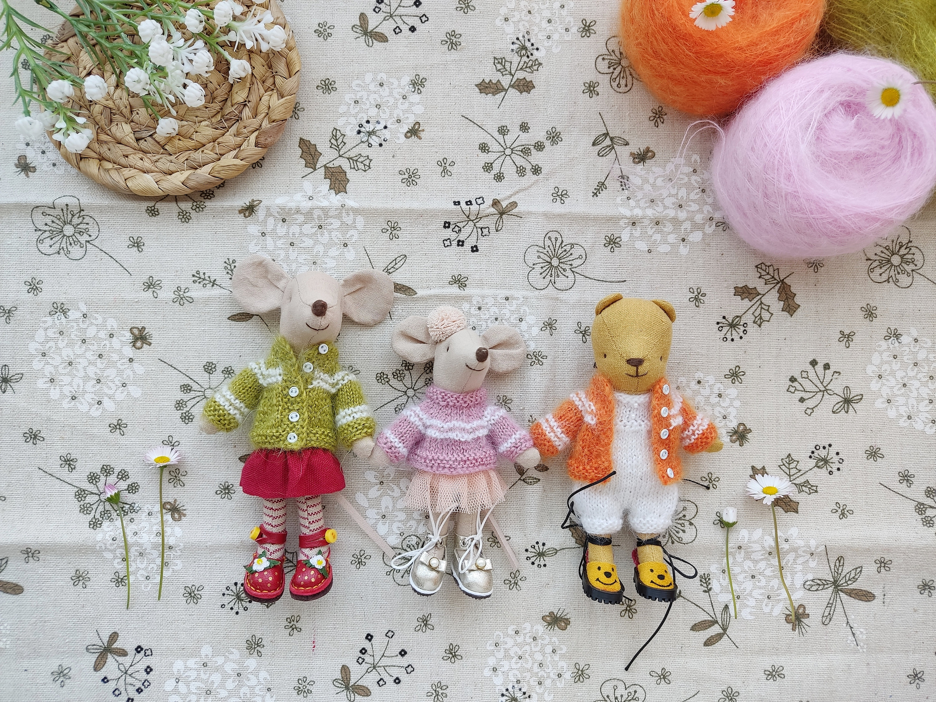 May include: Three handmade knitted toys: a green and white sweater-wearing mouse, a pink and white sweater-wearing mouse, and an orange and white sweater-wearing bear. The toys are sitting on a white fabric background with a floral pattern.