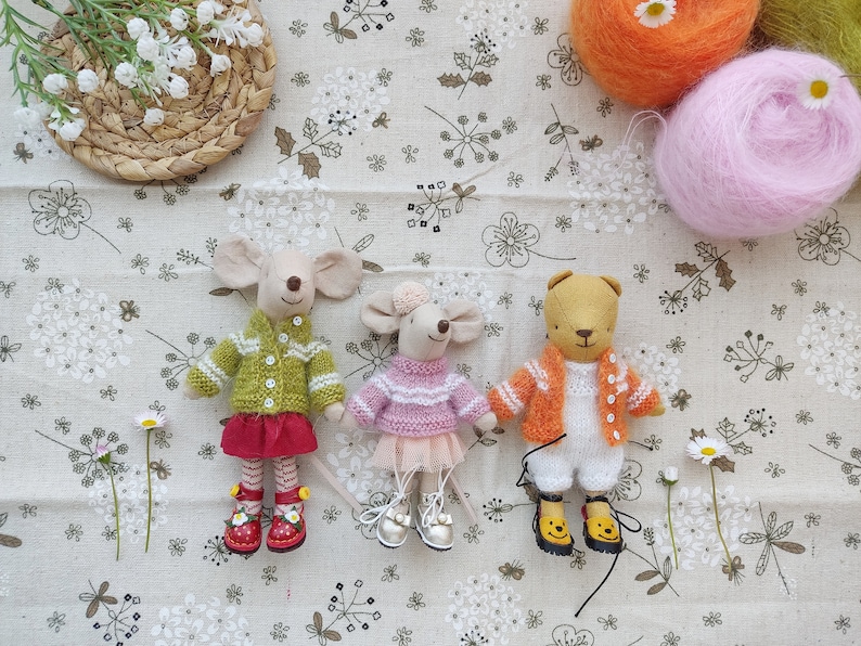 May include: Three handmade knitted toys: a green and white sweater-wearing mouse, a pink and white sweater-wearing mouse, and an orange and white sweater-wearing bear. The toys are sitting on a white fabric background with a floral pattern.