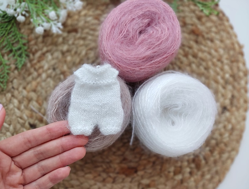 May include: A white knitted baby onesie with a pink and white yarn skein in the background.