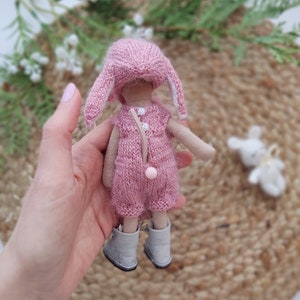 May include: A pink knitted doll with bunny ears, wearing a pink knitted dress and grey boots. The doll is being held by a hand.