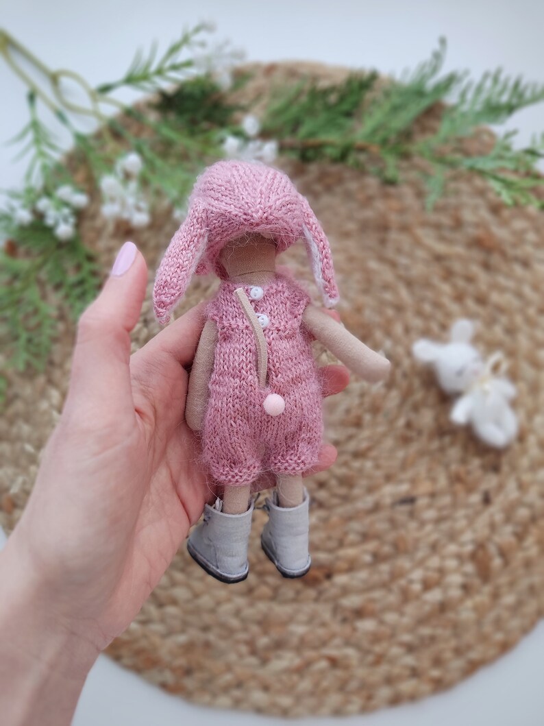 May include: A pink knitted doll with bunny ears, wearing a pink knitted dress and grey boots. The doll is being held by a hand.