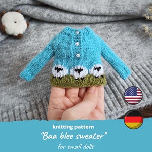 May include: A small, hand-knitted blue sweater with three sheep designs on the front. The sweater has long sleeves and three white buttons. The bottom hem is green. The text "Baa blee sweater" is visible.
