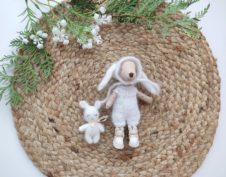 May include: Two white knitted stuffed animals, a bunny and a rabbit, on a woven jute rug. The rabbit has a white hood and a brown nose.