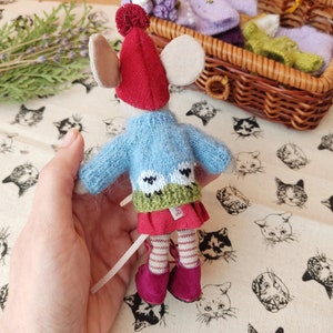 May include: A small knitted mouse doll wearing a red hat, a blue sweater with green and white accents, a red skirt, and red and white striped stockings. The doll is holding a white string in its hand.
