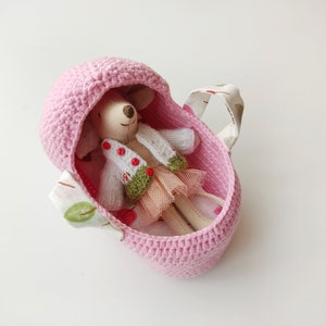 May include: A pink crocheted baby carrier with a fabric handle and a small doll inside wearing a white sweater and a green and pink skirt.