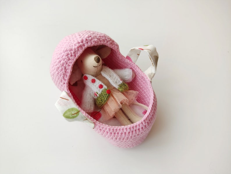 May include: A pink crocheted baby carrier with a fabric handle and a small doll inside wearing a white sweater and a green and pink skirt.