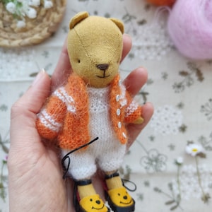 May include: A small, handmade plush bear wearing a white knitted onesie and an orange knitted cardigan. The bear has a brown nose and is wearing yellow shoes with a smiley face design.
