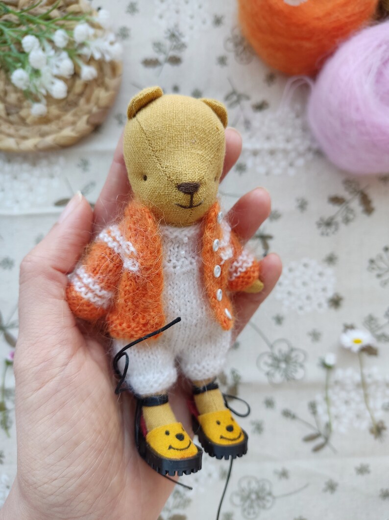 May include: A small, handmade plush bear wearing a white knitted onesie and an orange knitted cardigan. The bear has a brown nose and is wearing yellow shoes with a smiley face design.