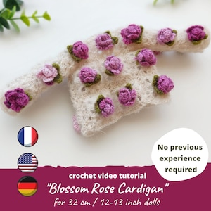 Puede incluir: Un cárdigan de crochet con base blanca y adornos de rosas rosas. El cárdigan está diseñado para muñecas de 32 cm. El texto "Blossom Rose Cardigan" y "tutorial de video de crochet" es visible en la imagen.