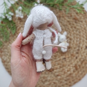 May include: A white knitted doll with bunny ears, wearing a white knitted outfit and white shoes. The doll is being held in a hand.