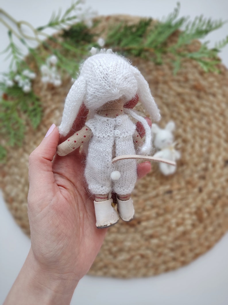 May include: A white knitted doll with bunny ears, wearing a white knitted outfit and white shoes. The doll is being held in a hand.