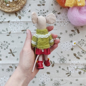 May include: A handmade knitted mouse doll wearing a green and white striped sweater and red skirt. The doll has red shoes and a long tail.