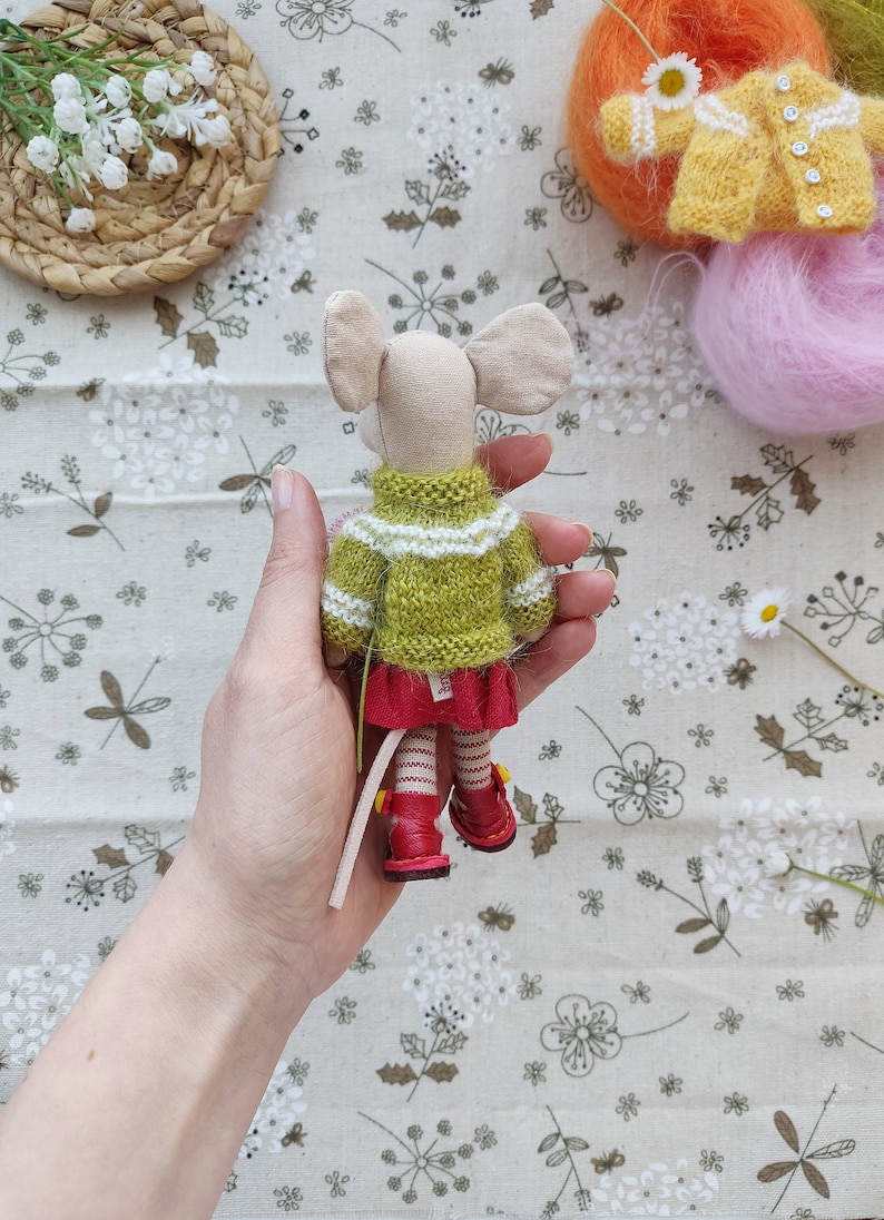May include: A handmade knitted mouse doll wearing a green and white striped sweater and red skirt. The doll has red shoes and a long tail.