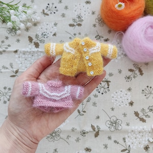 May include: Two knitted sweaters for dolls, one yellow with white trim and buttons, and one pink with white trim.