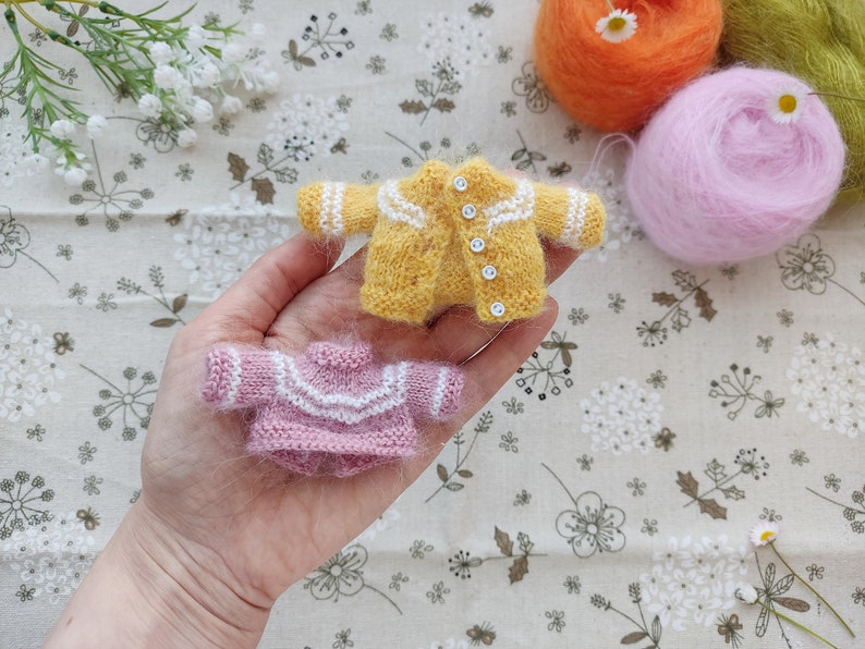 May include: Two knitted sweaters for dolls, one yellow with white trim and buttons, and one pink with white trim.
