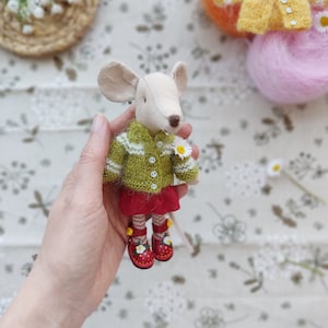 May include: A small, handmade mouse doll wearing a green knitted sweater with white trim, a red skirt, and red shoes with white flowers. The doll is being held by a hand against a white background with a floral pattern.