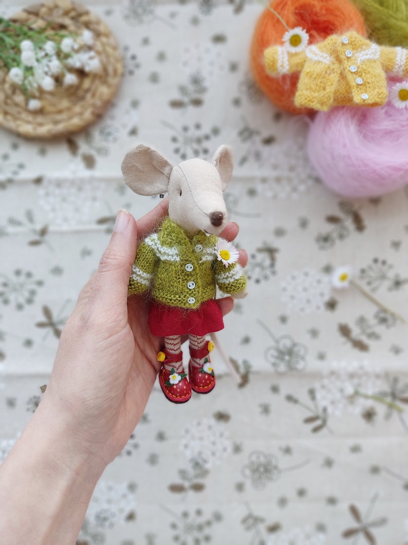 May include: A small, handmade mouse doll wearing a green knitted sweater with white trim, a red skirt, and red shoes with white flowers. The doll is being held by a hand against a white background with a floral pattern.