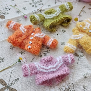 May include: Four miniature knitted sweaters in different colors: orange, green, yellow, and pink. Each sweater has white trim and buttons.