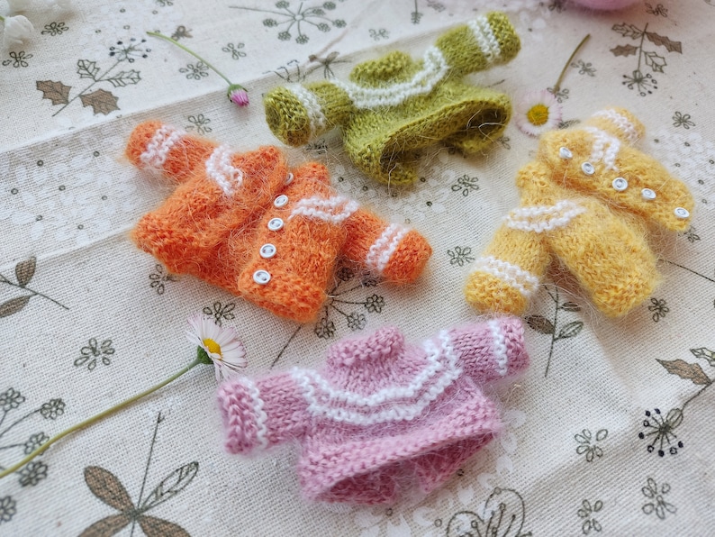 May include: Four miniature knitted sweaters in different colors: orange, green, yellow, and pink. Each sweater has white trim and buttons.