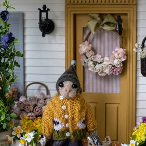 May include: A small, knitted animal wearing a yellow sweater and a grey hat stands in front of a miniature house with a wreath on the door. The animal is holding gardening tools and there are baskets and flowers in the scene.