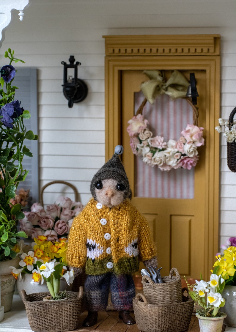 May include: A small, knitted animal wearing a yellow sweater and a grey hat stands in front of a miniature house with a wreath on the door. The animal is holding gardening tools and there are baskets and flowers in the scene.