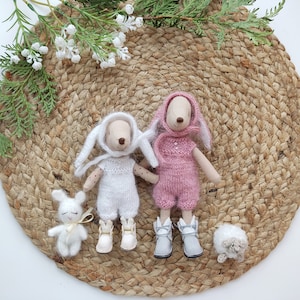 May include: Two plush bunny toys wearing knitted outfits. One bunny is wearing a white outfit and the other is wearing a pink outfit. The bunnies are sitting on a woven straw mat.