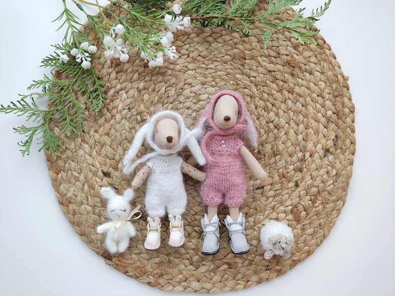May include: Two plush bunny toys wearing knitted outfits. One bunny is wearing a white outfit and the other is wearing a pink outfit. The bunnies are sitting on a woven straw mat.