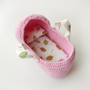 May include: A pink crocheted doll crib with a white and brown patterned fabric lining and a white pillow.