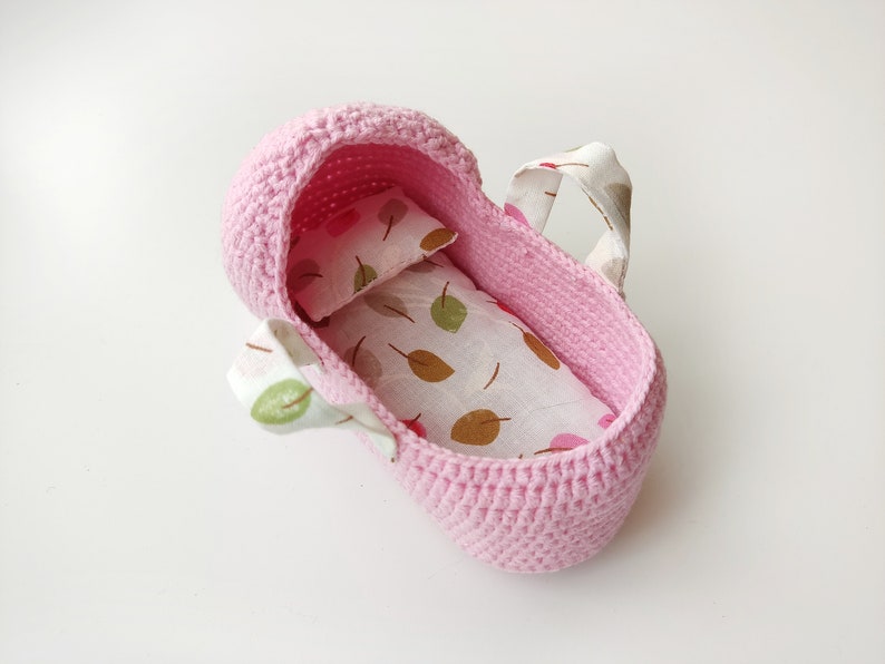 May include: A pink crocheted doll crib with a white and brown patterned fabric lining and a white pillow.
