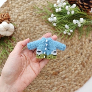 May include: A tiny blue knitted cardigan with white buttons and green trim. The cardigan has a sheep design on the front.