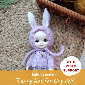 May include: Knitting pattern for a purple bunny hat for a tiny doll. The hat has white bunny ears and the doll is wearing a purple sweater with a bee pattern. The text "WITH VIDEO SUPPORT" is visible in a red circle.