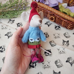 May include: A small, grey mouse doll wearing a blue jumper with a green and white sheep design, a red skirt, and red boots with white laces. The doll is being held in a hand.