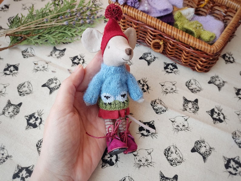May include: A small, grey mouse doll wearing a blue jumper with a green and white sheep design, a red skirt, and red boots with white laces. The doll is being held in a hand.