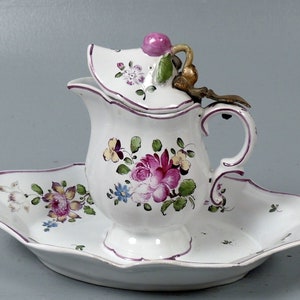 Puede incluir: Una jarra de porcelana blanca con un diseño floral y una tapa, sentada sobre un plato de porcelana blanca a juego con un borde festoneado.