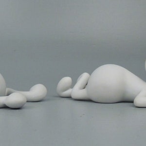 Può includere: Due sculture in ceramica bianca di figure astratte. Una figura è seduta con le gambe incrociate, mentre l'altra è sdraiata di lato con la testa appoggiata sulla mano.