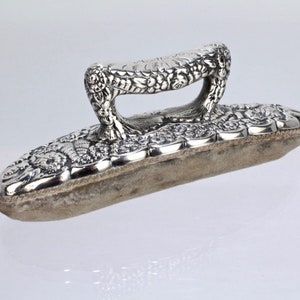 May include: A silver-toned, rectangular shoe shine brush with an ornate handle. The handle and the brush's frame feature a floral design. The brush has a light-colored, rectangular pad for applying shoe polish.