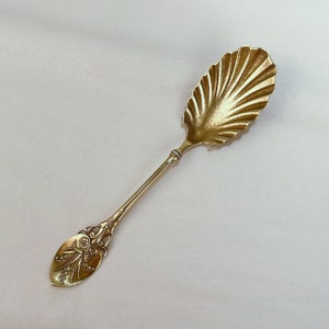 May include: A gold-colored sugar spoon with a decorative, scalloped bowl and a floral design on the handle.