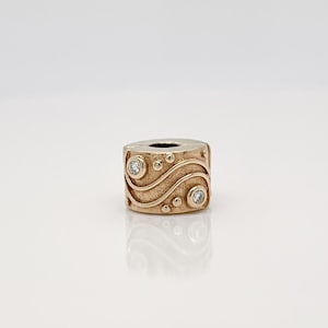 Puede incluir: Encanto de oro con un diseño de espiral y dos pequeños diamantes transparentes. El encanto tiene forma cilíndrica y un acabado liso y pulido.
