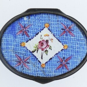 Könnte beinhalten: Eine ovale, silberfarbene Schmuckdose mit einem blauen und weißen Emaille-Design. Der Deckel zeigt eine weiße Diamantform mit einer rosa Rose und grünen Blättern in der Mitte. Der Diamant ist von einem blauen Gittermuster mit vier rosa und violetten sternförmigen Blumen umgeben.