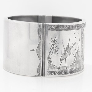 Puede incluir: Un brazalete de bisagra de plata con un dise&ntilde;o grabado de un p&aacute;jaro en un entorno de pantano.