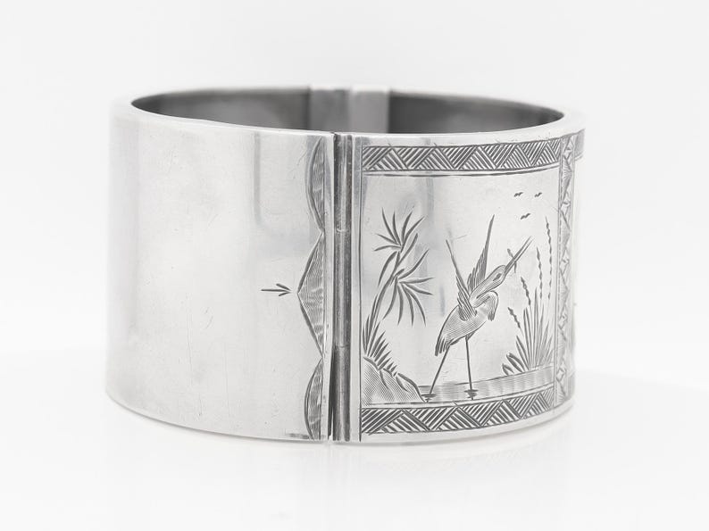 Puede incluir: Un brazalete de bisagra de plata con un dise&ntilde;o grabado de un p&aacute;jaro en un entorno de pantano.