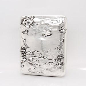 Puede incluir: Un estuche de cerillas rectangular de plata con tapa abisagrada. La parte delantera del estuche de cerillas presenta un diseño en relieve detallado de peces nadando en el agua, rodeados de plantas acuáticas. Los bordes y la parte superior también están decorados con patrones texturizados.
