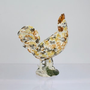 Puede incluir: Una figura de gallo de cerámica con una base blanca y un patrón moteado marrón, amarillo y negro. El gallo tiene una cresta y barbillas rojas.