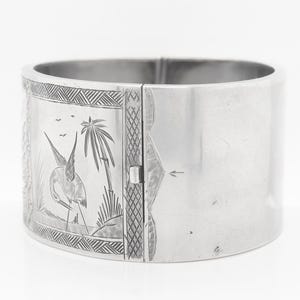 Puede incluir: Un brazalete de plata con un dise&ntilde;o grabado de un p&aacute;jaro y palmeras. El brazalete tiene bisagras y se abre para ajustarse a la mu&ntilde;eca.