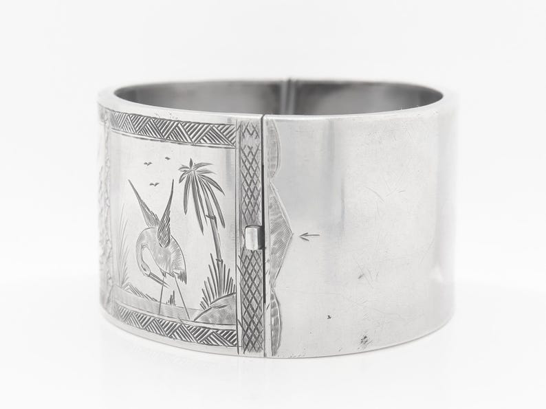 Puede incluir: Un brazalete de plata con un dise&ntilde;o grabado de un p&aacute;jaro y palmeras. El brazalete tiene bisagras y se abre para ajustarse a la mu&ntilde;eca.