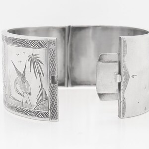 Puede incluir: Un brazalete de bisagra de plata con un dise&ntilde;o grabado de un p&aacute;jaro y palmeras.