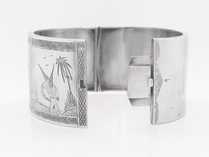 Puede incluir: Un brazalete de bisagra de plata con un dise&ntilde;o grabado de un p&aacute;jaro y palmeras.