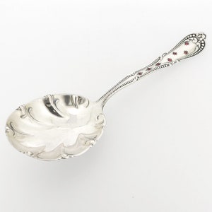May include: A silver serving spoon with a decorative handle featuring red enamel accents. The bowl of the spoon is scalloped and has a swirl design.
