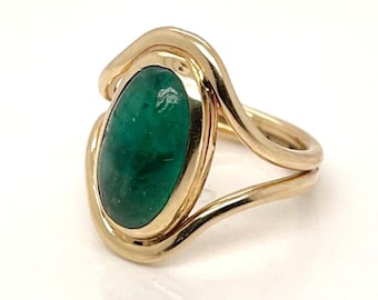 May include: A gold ring with a large, oval-shaped green gemstone set in a bezel setting. The ring band is a double band that wraps around the gemstone.