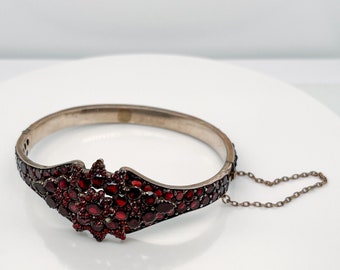 May include: A silver bangle bracelet with a decorative design of red garnets. The bracelet has a chain attached to the clasp.